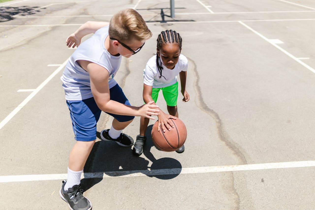 Two children playing basketball outdoors on a sunny day, practicing their dribbling skills.